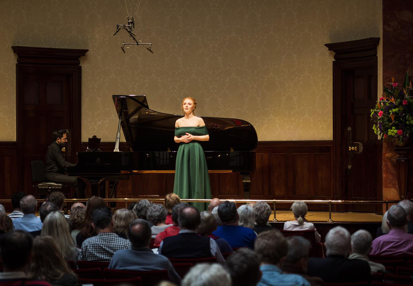 Esme Bronwen-Smith singing on stage to an audience in the foreground, and an accompanist is playing the piano behind her